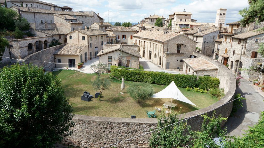 Vista dall'alto dell'Anfiteatro Romano di Assisi con un giardino verde e le mura che racchiudono alcune case di cittadini della città. Foto scattata di giorno con luce solare.