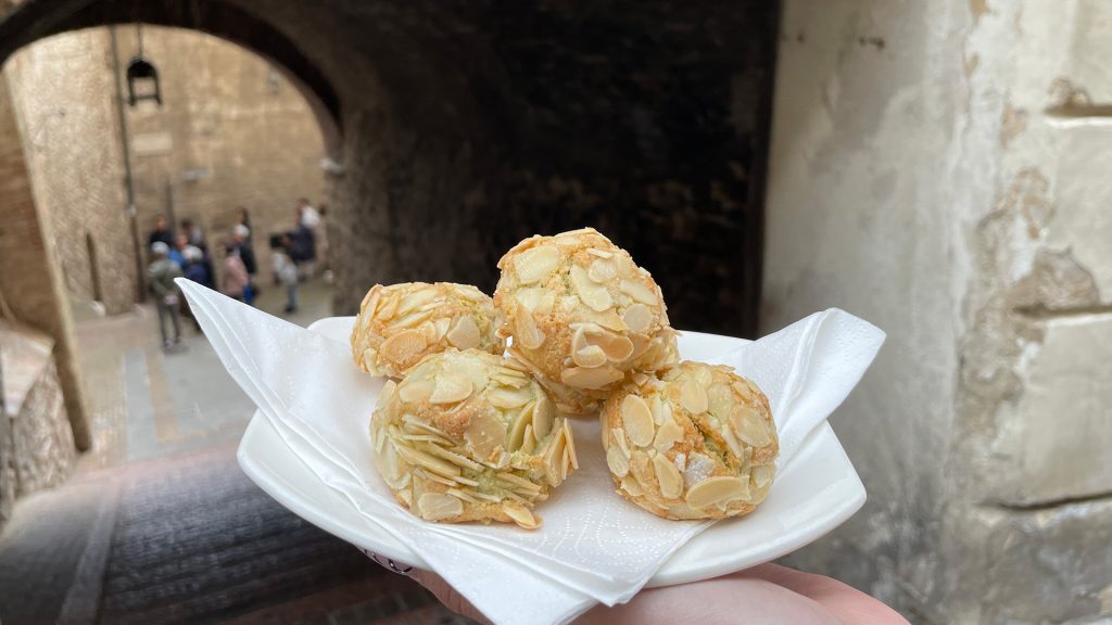 Nella foto è presente uno scorcio sul centro storico ed in primo piano è presente un vassoio con al suo interno il tipico dolce "Il bacio di Assisi". Il docle è composta da una sfoglia. Hanno una forma a pallina con sopra zucchero, pistacchio o altro.