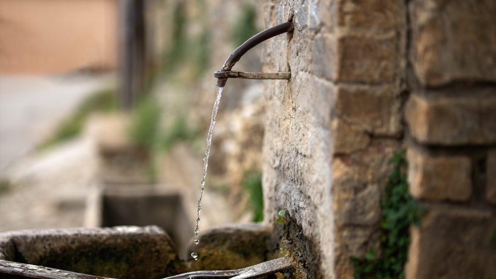 Vista di una fontana pubblica della città di Assisi. Un dettaglio della fontanina con un tubo in rame da cui esce l'acqua e finisce nella sua vasca di pietra. La fontanina è sorretta da un anello in pietra.