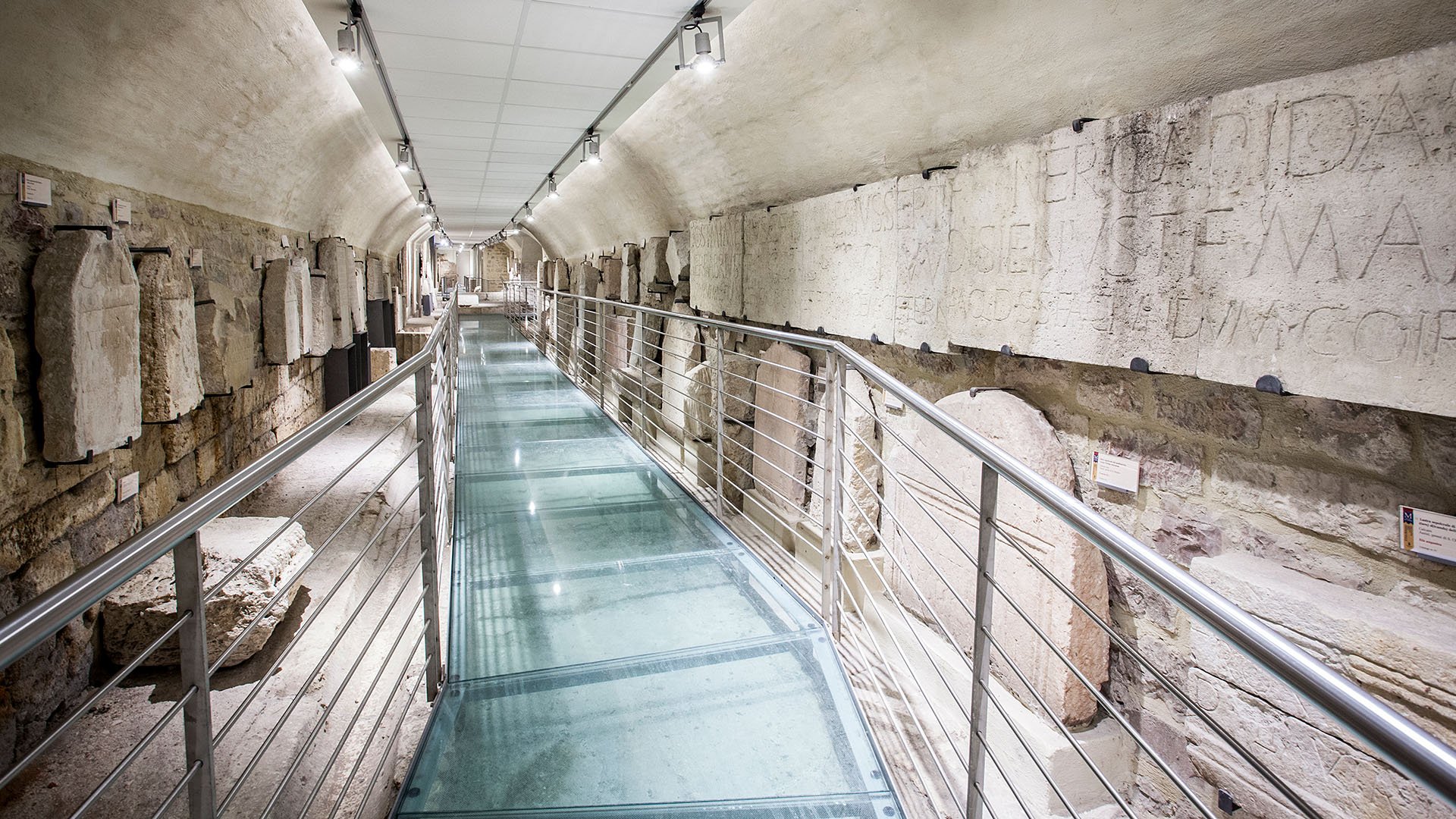 Vista interna del corridoio del museo del Foro Romano con ai lati tutti sassi e pietre con scritte dell'epoca romana ed al centro la passerella con pavimentazione in legno ed corrimano laterali in ferro.