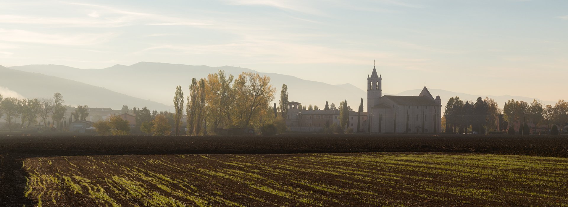 Vista da lontano e laterale del Santuario Francescano di Rivotorto, con il campanile che svetta al centro e laterale il santuario. In primo piano invece un campo coltivato, la foto ha un'atmosfera suggestiva.