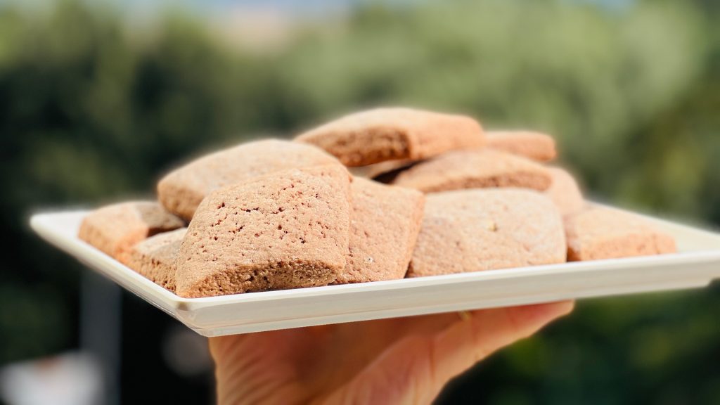 La foto rappresenta dei tipici dolci umbri chiamati Mostaccioli. Questi sono messi su un vassoio che sta venendo trasportato. Sullo sfondo si intravede del verde completamente sfuocato.