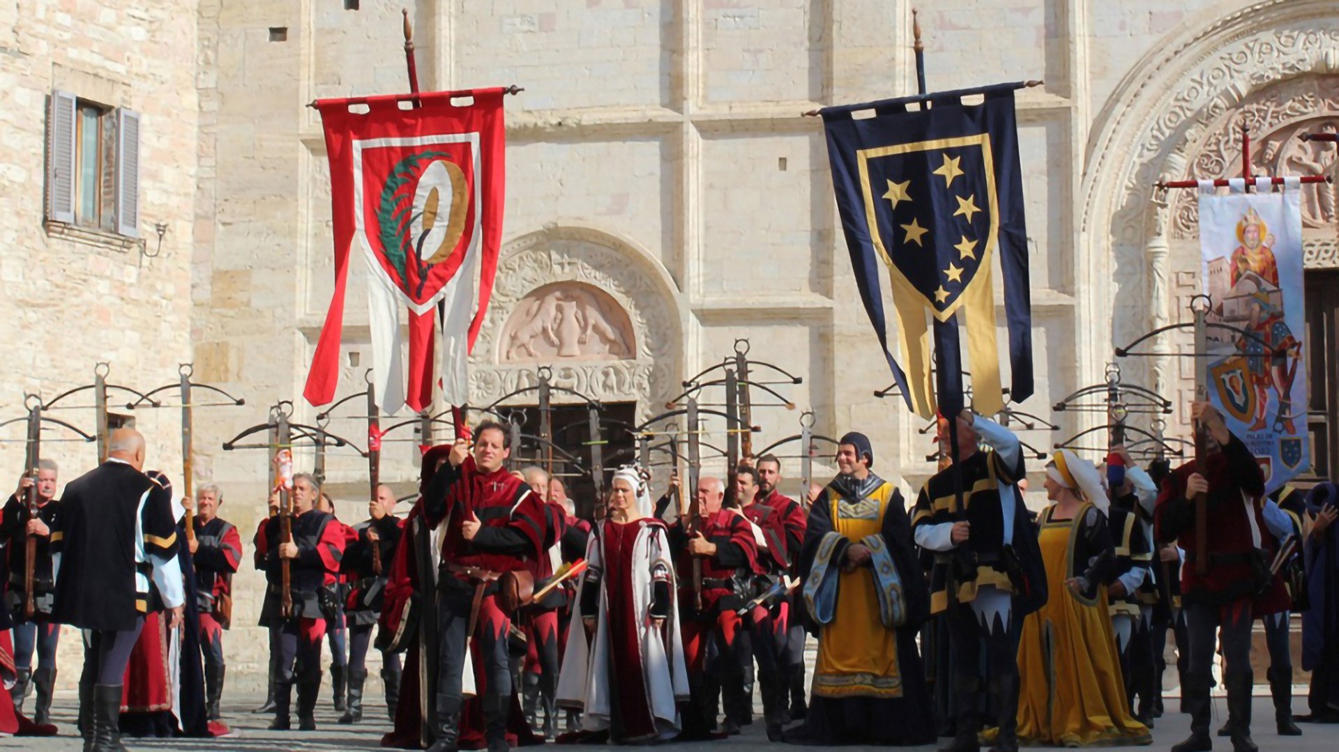 Foto che rappresenta una sfilata storica tipica del Palio di San Rufino. Nella foto possiamo vedere tutte persone vestite come in epoca medievale divisi in due file, è presente un capo fila che porta con se lo stendardo del suo rione.