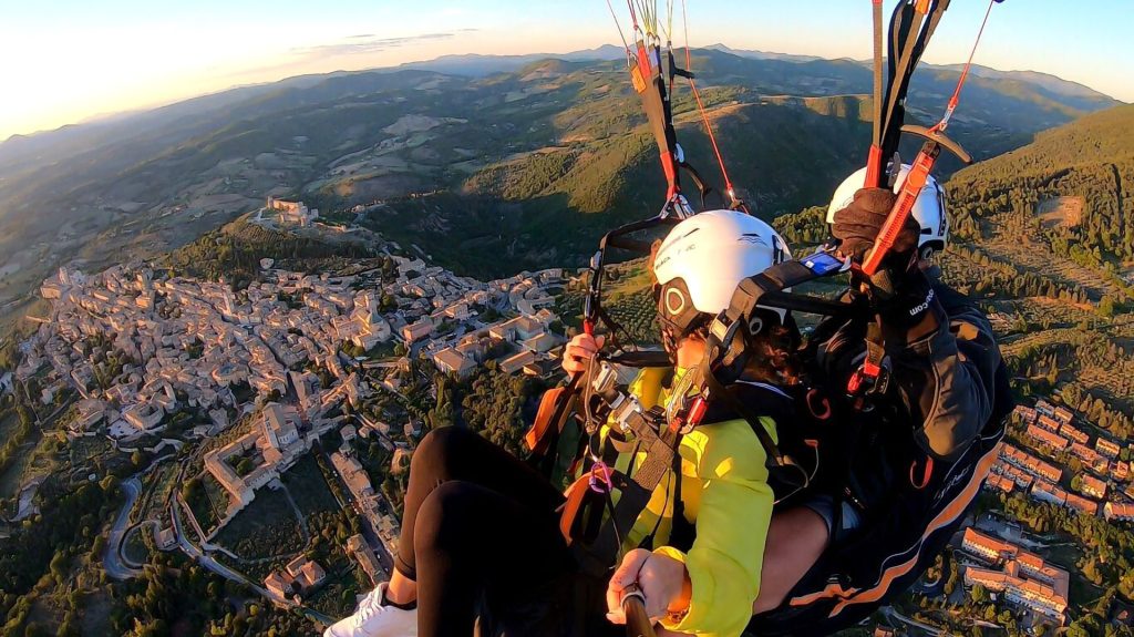 La foto riprende due persone in primo piano che sorvolano la città di Assisi effettuando del parapendio. In sfondo, sotto di loro, una bellisima vista dall'alto della città di Assisi.