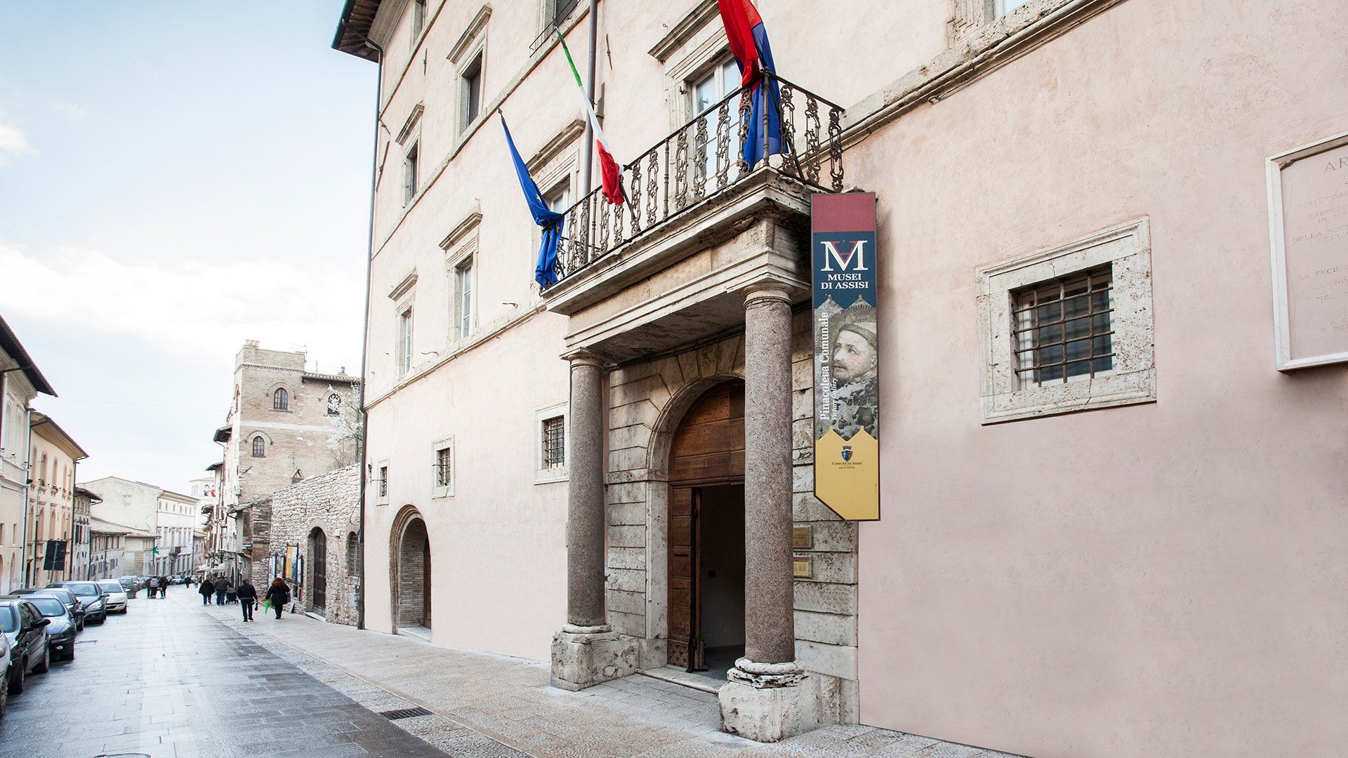 Immagine raggigurante la via dove è situato il museo della Pinacoteca comunale di Assisi. In primo piano abbiamo uno stendardo con la locandina del museo, alla sua sinistra abbiamo l'ingresso con le due colonne che sorreggono un balconcino dove sono presenti le bandiere dell'Italia e dell'Europa.