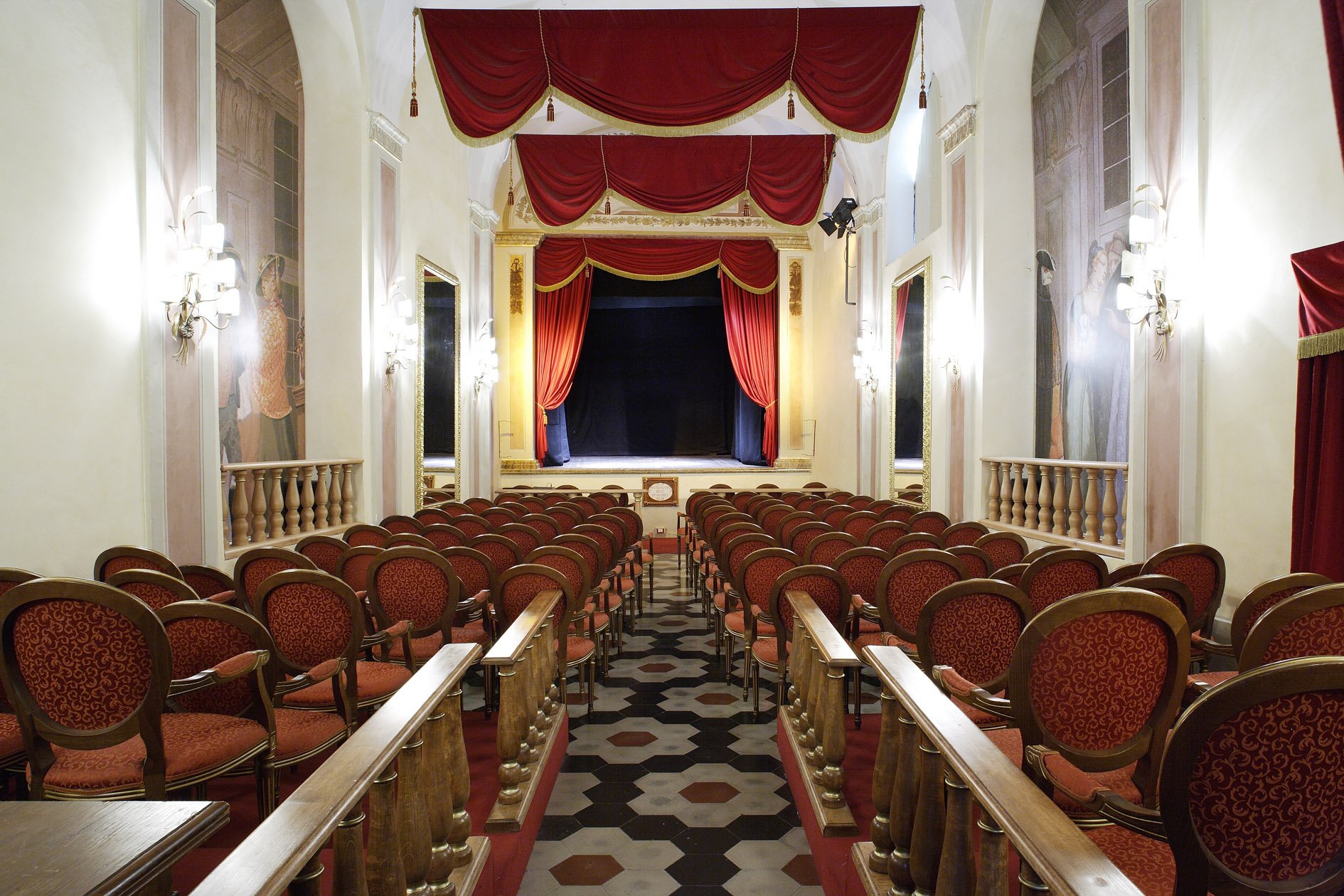 Immagine che rappresenta il piccolo Teatro degli instabili di Assisi. Il palco è in marmo bianco ed ai lati del palco le tende sono rosse con drappeggi in oro. Le poltrone sono in legno con seduta e schienale rosso con decorazioni floreali in ora. Il pavimento ha una texture esagonale bianca ed al centro un esagono arancione, il fondo del pavimento è scuro.